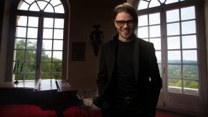 Portrait of pianist Grigorii Palamarchuk standing next to a grand piano in front of large arched windows.