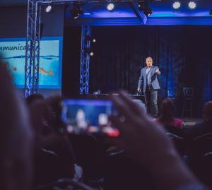 Dr. Dennis Cummins delivering a keynote presentation on stage, photographed from the audience perspective.