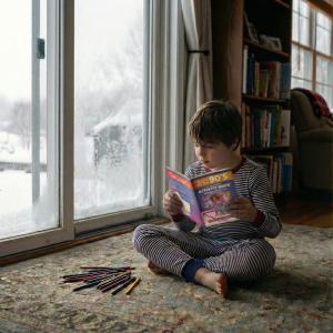 A child in striped pajamas sits on a rug by a large window with snow outside, holding and reading a colorful 90s activity book, with pencils scattered on the floor nearby.