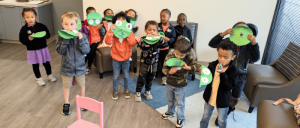 A group of children holding up green paper masks with eyes and mouths, smiling as they pose together in a brightly lit room.