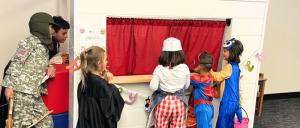Children in costumes, including a chef and superheroes, gather around a playhouse with a red curtain, engaging in imaginative play.