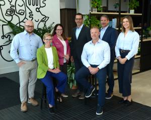 Denver-based consultancy Karrikins Group has been named to Inc Seven Karrikins Group team members pose for a professional group photo in a modern office with plants and abstract line art, with two seated on stools in front and five standing behind, all in business attire smiling at the camera, from left to right Dani