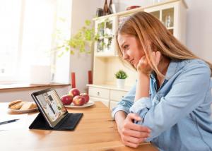 Woman smiling during a virtual telehealth consultation on tablet at home kitchen table, representing accessible mental health care services.