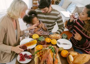 Multi-generational family sharing a holiday meal together around a table filled with traditional foods, with young child being fed while seated between family members.