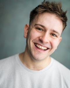 Max Cadman smiles warmly at the camera against a soft, blurred gray background. He wears a light grey sweater, conveying a joyful tone.