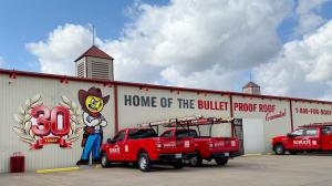 Schulte Roofing headquarters with red service trucks parked outside, a large BulletProof Bob mascot mural, and a ‘30 Years’ anniversary emblem on the building reading ‘Home of the BulletProof Roof Guarantee.’