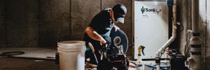 A TerraFirma foundation specialist in a dark uniform and cap kneels in a concrete basement next to a white SaniDry XP dehumidifier and a tool bag, preparing equipment for moisture control.