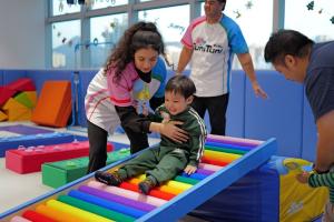 A young toddler in a green tracksuit slides down a colorful rainbow roller ramp, supported by a TuniTuni instructor, inside a bright play gym located in Hong Kong.