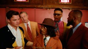 Ashwin Gane dressed as Inspector Gane in a cinematic still from the “Flip Dat” music video, standing in the center with five characters reacting dramatically around him.