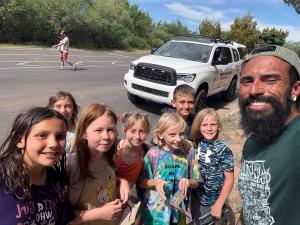 A group of smiling children posing with an outdoor guide in front of a white off-road vehicle during a community nature outing.