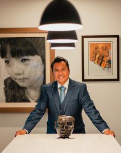 Johnessco Rodriguez, directeur d’ARQA ART FAIR, debout aux côtés d’œuvres de sa collection personnelle, dont un objet précolombien de Teotihuacan, avec des œuvres encadrées visibles en arrière-plan.