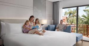 A family sitting on a large bed and lounge chair in a modern hotel room with a balcony overlooking palm trees at Courtyard by Marriott Phuket, Patong Beach Resort.