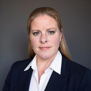 Professional headshot of a woman wearing a navy blazer and white collared shirt, facing the camera against a neutral gray background.