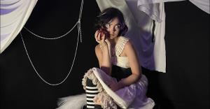 Alisha Liston Announces the Expansion of Dreamland with her Newest Release Angelology 10 Alisha Liston sitting on set in a purple skirt, white blouse and black and white stockings holding a pomegranate in her hand