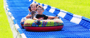 Two children enjoy a colorful inner tube as they slide down a bright blue water slide on a sunny day, surrounded by green grass.