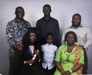 A family picture delebrating Christabel's graduation. Christabel sits in on a chair wearing her graduation outfit holding her certifciate. A young boy sits next to her besid her mother. Behind her is her father with his hand on her shoulder and two other 