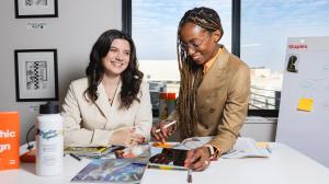 Two women professionals, Nile Livingston standing beside Tara Rose O’Connor during a collaborative conversation in a Creative Repute office, with design tools, notebooks, and visual references on the table.