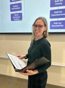 A woman holding an open binder smiles while standing in front of a projected screen.
