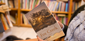 A person holding a copy of the novel Fox Creek by M. E. Torrey in a library setting, with bookshelves in the background.