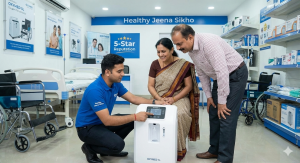Two professionals standing in a medical equipment showroom shaking hands over an OXYMED oxygen concentrator. The man on the left wears a corporate navy blazer, while the man on the right wears a light blue medical uniform with the 'Healthy Jeena Sikho' lo