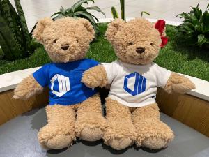 Close-up of the Hope and Hugo mascots sitting on a grey bench in a modern office lobby surrounded by green indoor plants.