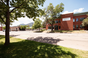 Exterior shot of Strive Boulder. Image features red brick building against a backdrop of mountains. Green courtyard and trees in the foreground