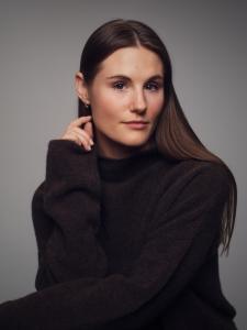 Professional studio portrait of a female actor with brown hair and blue eyes, wearing a dark long-sleeve top, photographed against a neutral gray background.