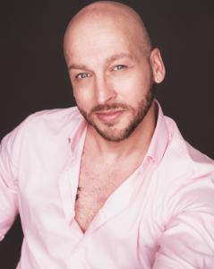 Professional studio headshot of a male actor with a shaved head and short beard, wearing a light pink button-down shirt, photographed against a dark neutral background.