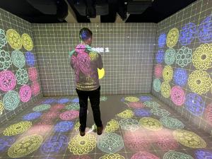 A visitor interacts with immersive visuals projected across the floor and walls inside NEWJAK’s six-sided XR space. The experience requires no wearable devices, relying solely on physical movement.