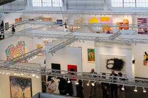 Interior view of the San Francisco Art Fair at Fort Mason Festival Pavilion with gallery booths, contemporary artworks on display, overhead lighting trusses, and visitors exploring the exhibition space.