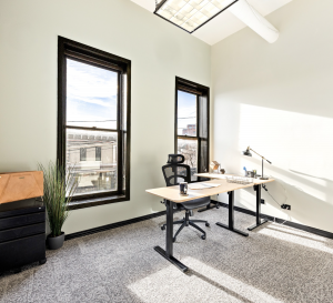 Image features a mid-sized exterior private office, with white walls, natural wood desk and black ergonomic desk chair