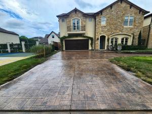 A wide, freshly sealed stamped-concrete driveway leads to a two-story stone-and-stucco house with a dark brown double garage, under a cloudy sky.