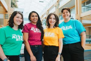 Photo shows four people wearing colourful t-shirts that read, UNBC Active Minds. The group is standing in a building atrium with banners displaying Indigenous artwork in the background.