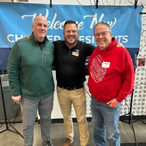 Three people pose together smiling at a Sleep in Heavenly Peace chapter training event, standing in front of a blue banner reading "Welcome New Chapter Presidents." The person in the center wears an SHP polo shirt, flanked by two volunteer chapter leaders