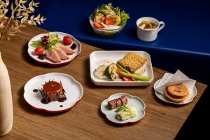 A plated inflight meal set displayed on a wooden table, featuring oat-crusted cod with vegetables and sauce, green papaya salad with scallop, assorted appetizers including stuffed squid and beef roll, a dessert, and a wheel pie.