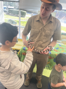 Educator holds a snake while a child gently touches it near a window inside a center.