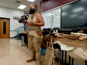 A man dressed as a caveman teaches a class about the Ice Age.  He is holding a fossil sabertooth cat skull in front of a display of prehistoric Stone Age replicas.