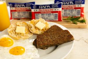 Plated RAPA Scrapple with sunny-side-up eggs and an English muffin, with RAPA Scrapple packages in the background.