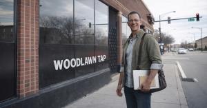 Chris J. Parker smiling on the sidewalk outside the Woodlawn Tap (Jimmy’s) in Chicago, holding a book and bag ahead of his book signing.