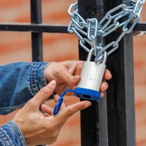 A female hand using a fingerprint to unlock a Matte Royal Blue BenjiLock padlock secured to an outdoor gate. The stainless steel surface of the lock is covered in water droplets, demonstrating its IP68 waterproof and weather-resistant capabilities in wet 
