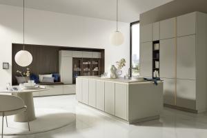 An ultra-modern, open-concept kitchen in a soft greige color. It features a central island and a recessed wall unit with a dark wood bench and blue pillows. Polished white marble floors reflect the soft glow of globe pendant lights.