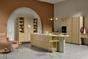 A bright kitchen with vertical-grain light oak cabinetry. A long island with a white countertop is paired with a yellow wire-frame barstool. A large terracotta-colored arch on the back wall serves as a backdrop for a display cabinet.