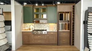 A kitchen display featuring forest green upper cabinets and warm brown wood lower cabinets with a herringbone-patterned tile backsplash.