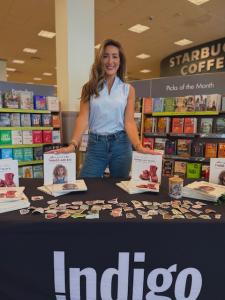 Shelby Moore standing behind a signing table at an Indigo bookstore, displaying copies of When You’re Little, Things Are Scary and When You’re Little, Things Are Big, with children’s bookshelves visible in the background.