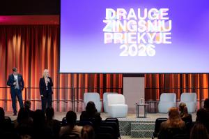 Du pranešėjai – vyras ir moteris – stovi konferencijos scenoje priešais didelį ekraną su užrašu „Drauge žingsniu priekyje 2026“. Fone – oranžinės užuolaidos, scenoje pastatytos pilkos kėdės ir staliukas, o pirmame plane matyti sėdinti auditorija.