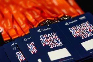 Tamsiai mėlyni renginio ženkleliai su užrašu „Drauge žingsniu priekyje 2026“ ir „Peoplelink“ logotipu sudėti kartu su ryškiai oranžinėmis kaklajuostėmis.