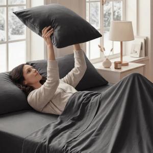 Beddora introduces its 4-Piece Sheet Set, offering Canadians premium comfort, long-lasting durability, and versatile design for any bedroom Woman lying on a bed covered with dark gray silky sheets, holding a matching pillow in a bright, modern bedroom with a bedside table and lamp.