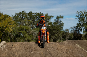 Gage Linville sits on an orange #74 KTM dirt bike during a team photoshoot at an outdoor dirt track, wearing orange and black race gear with trees and blue sky behind him.