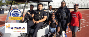 Two police officers sit in a golf cart while a child stands behind them, smiling. A track and field area is visible in the background.