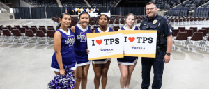 A group of cheerleaders in uniforms and a police officer hold signs reading 